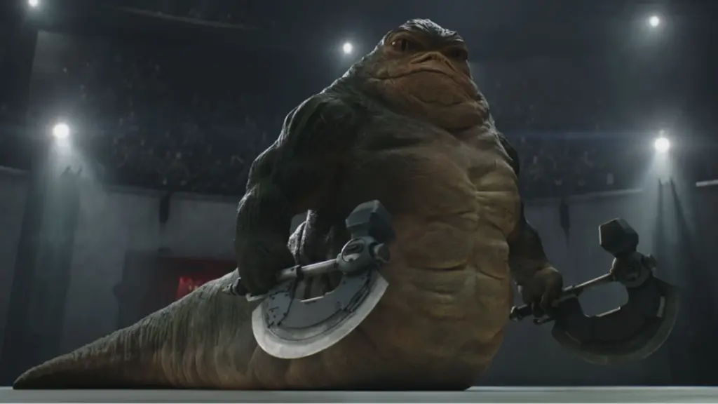Rotta the Hutt, voiced by Jeremy Allen White, shows off his buff muscles while holding two battle axes in the middle of a giant gladiator pit in THE MANDALORIAN AND GROGU. 
