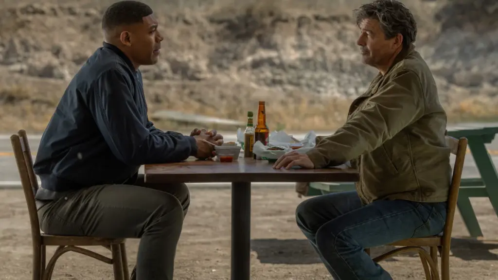 Aaron Pierre and Kyle Chandler star as John Stewart and Hal Jordan sitting together at an outdoor restaurant with a desert backdrop in HBO's LANTERNS.