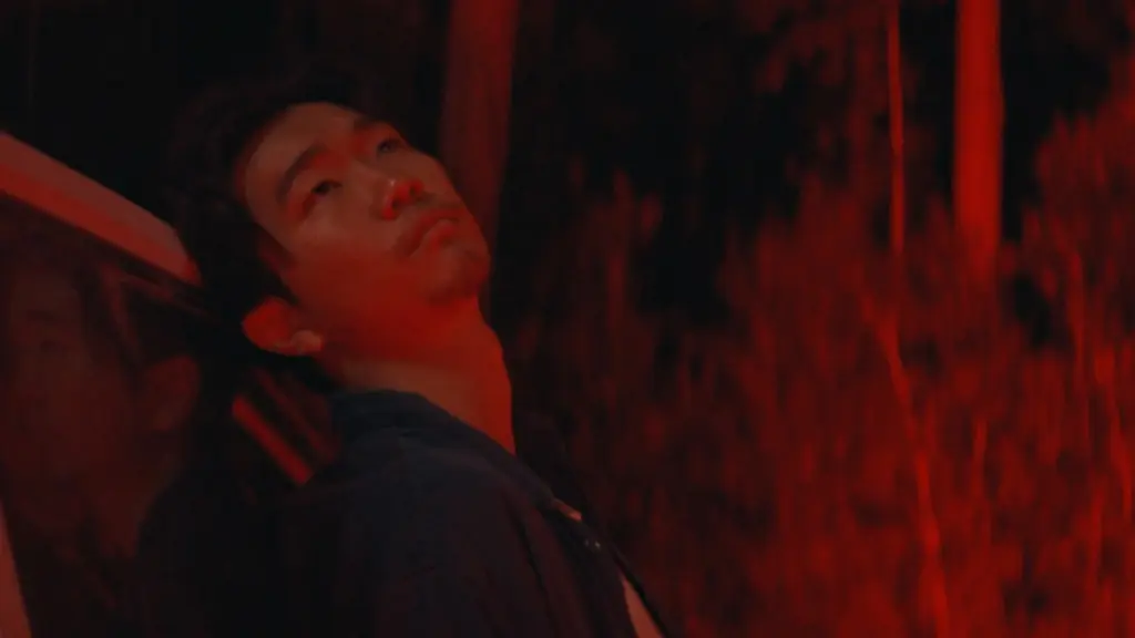 Phinehas Yoon as the character James looks up in despair while leaning against a car and being lit by a neon red light in the horror film IT ENDS.  