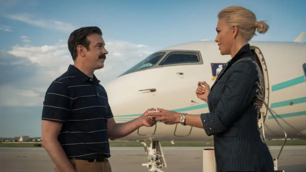 Jason Sudeikis and Hannah Waddingham firmly shake hands in front of a large jet in TED LASSO Season 4. 