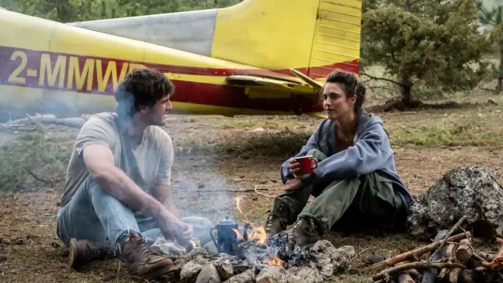 Jacob Elordi and Margaret Qualley sit by a camp fire next to a small yellow 1956 Cessna 182 plane in THE DOG STARS. 